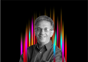 On the middle of a black background is a row of colourful vertical stripes’. In the centre is a black and white profile photo of TEDx speaker Sir Julian Le Grand superimposed on top.